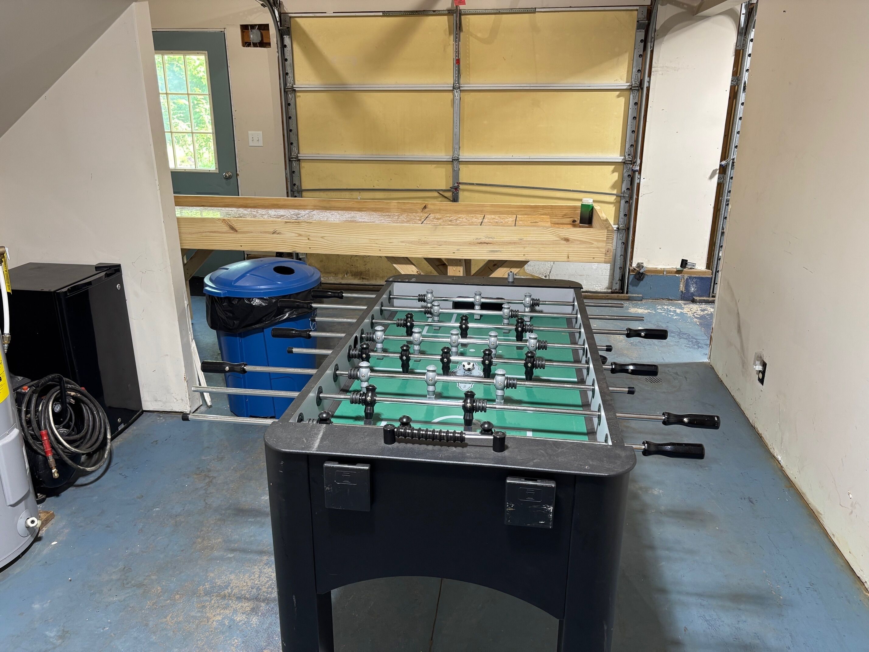 Een tafelvoetbaltafel in een ruimte met een garagedeur, een blauwe prullenbak en een zwarte speaker.