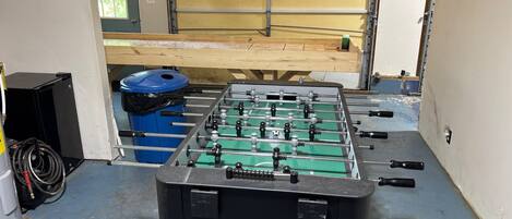 Een tafelvoetbaltafel in een ruimte met een garagedeur, een blauwe prullenbak en een zwarte speaker.