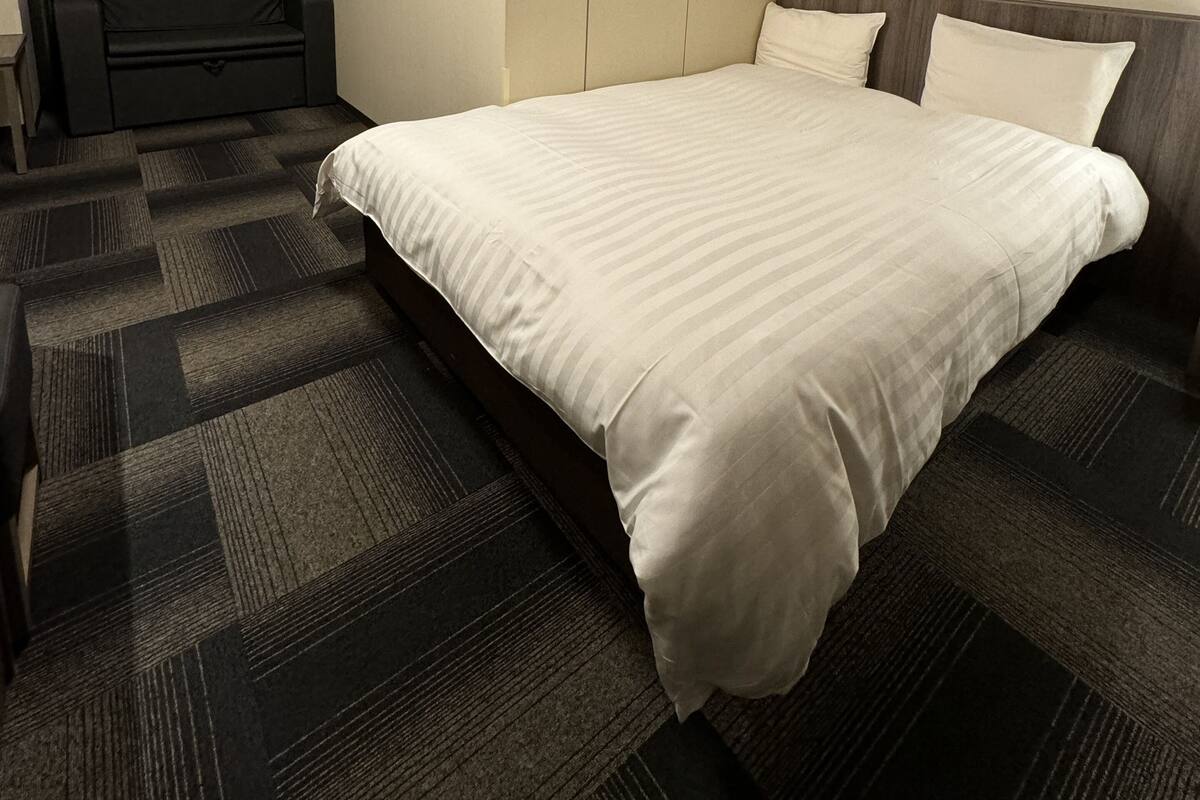 Une chambre d’hôtel avec un grand lit, un fauteuil sombre et un mur uni.