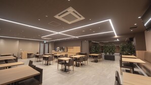 A modern dining area with multiple tables and chairs, a kitchen area, and a seating area with plants. - Dormy Inn Korakuen Hot Springs (Tokyo)