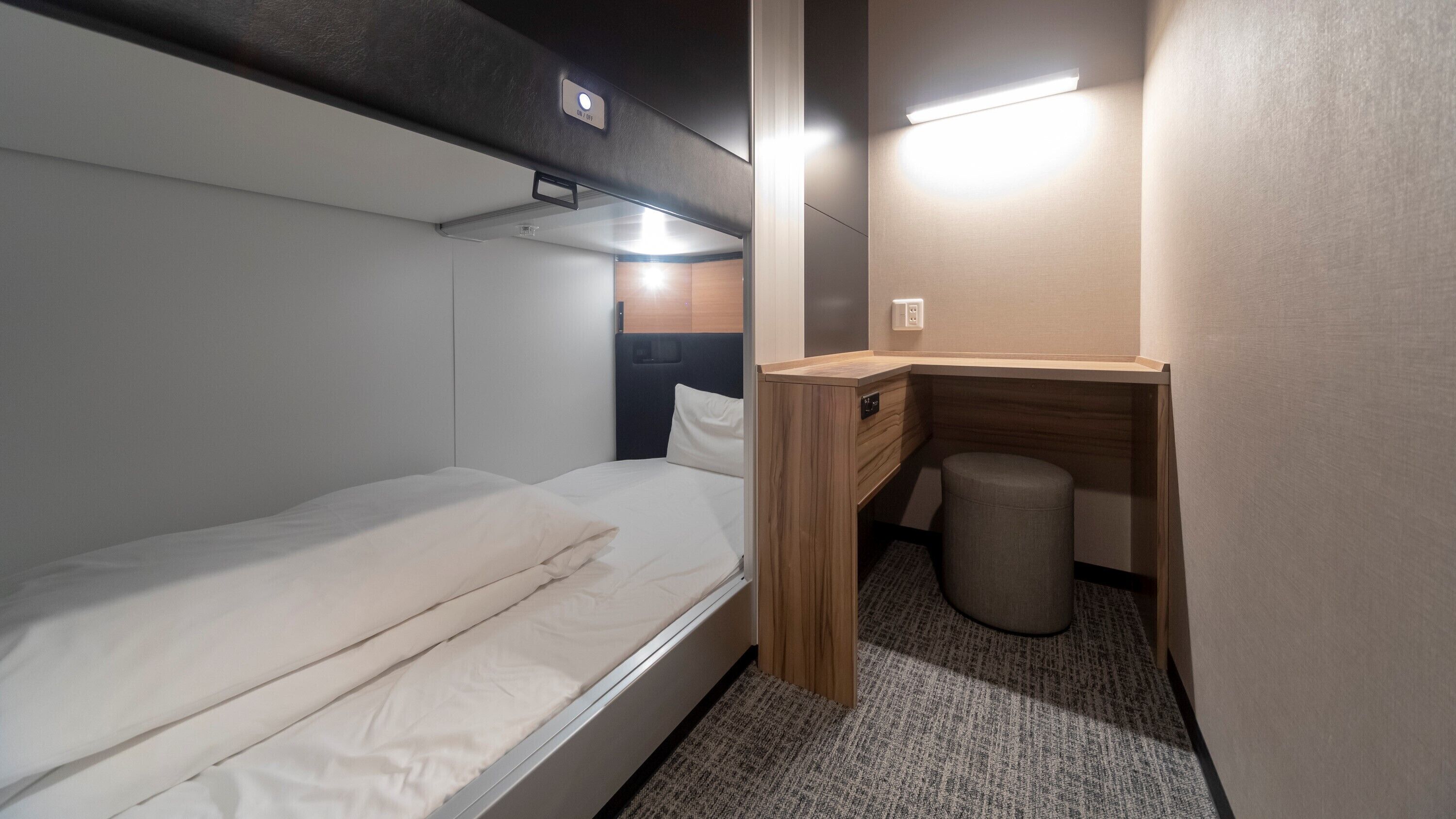A compact bedroom with a single bed, a small desk, a waste bin, and a wall-mounted light.