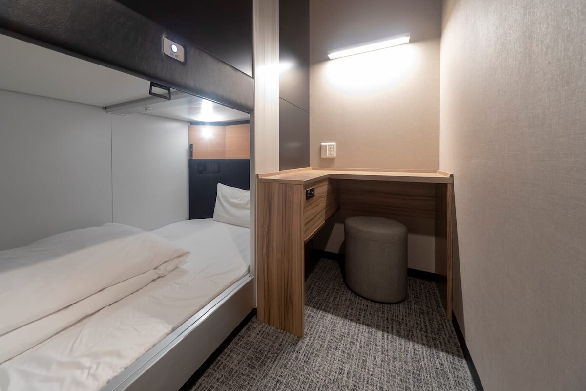 Une chambre à coucher de taille réduite, comprenant un lit simple, un petit bureau, une poubelle et une lampe fixée au mur.