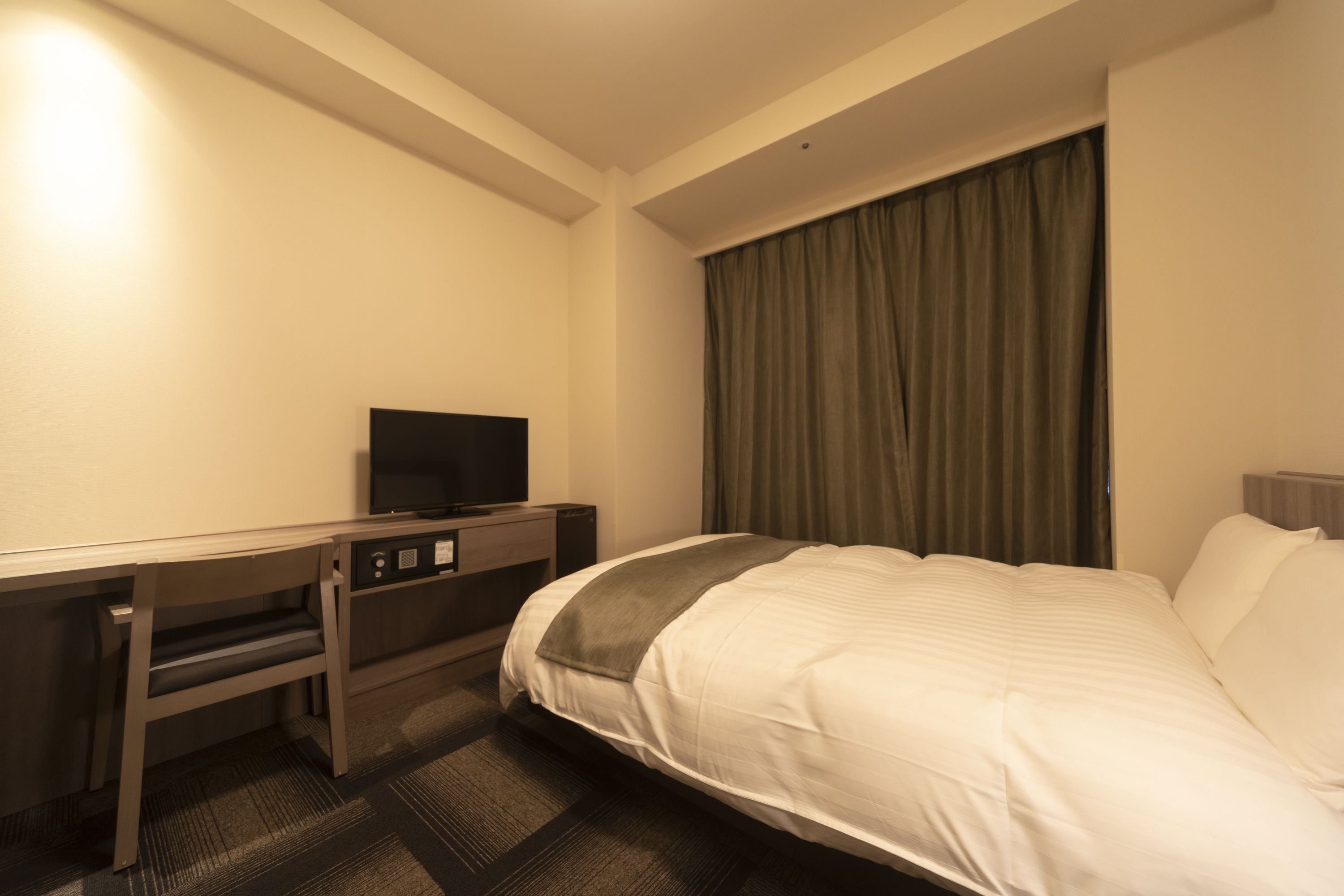 A hotel room with a bed, a desk with a television, and a window with curtains.