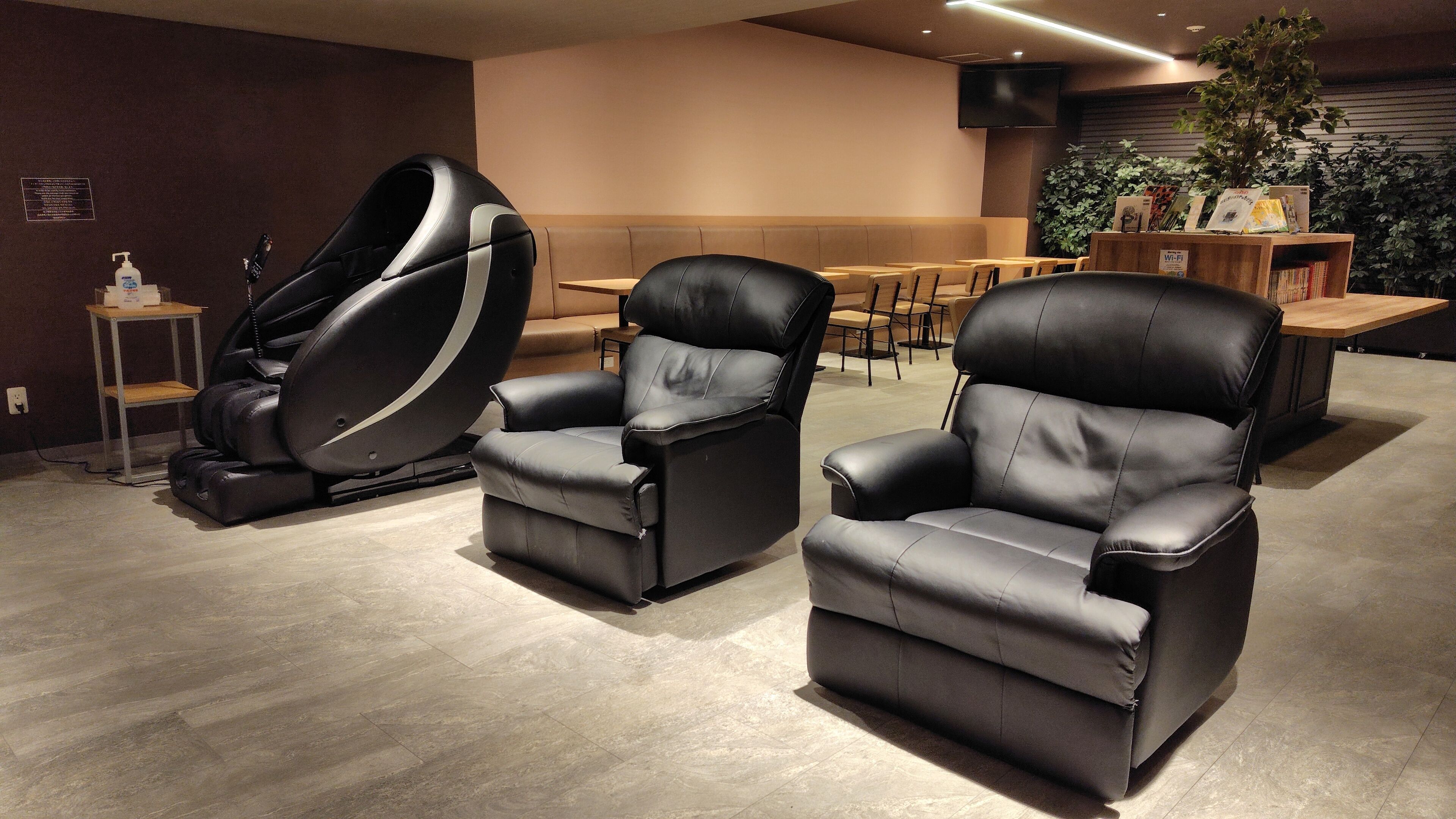A massage chair area with two massage chairs and a foot massage chair, a small table with a bottle, and a bookshelf with books and a plant.