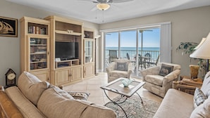 A spacious living room with a large TV, comfortable seating, a ceiling fan, and a balcony with a view of the ocean.