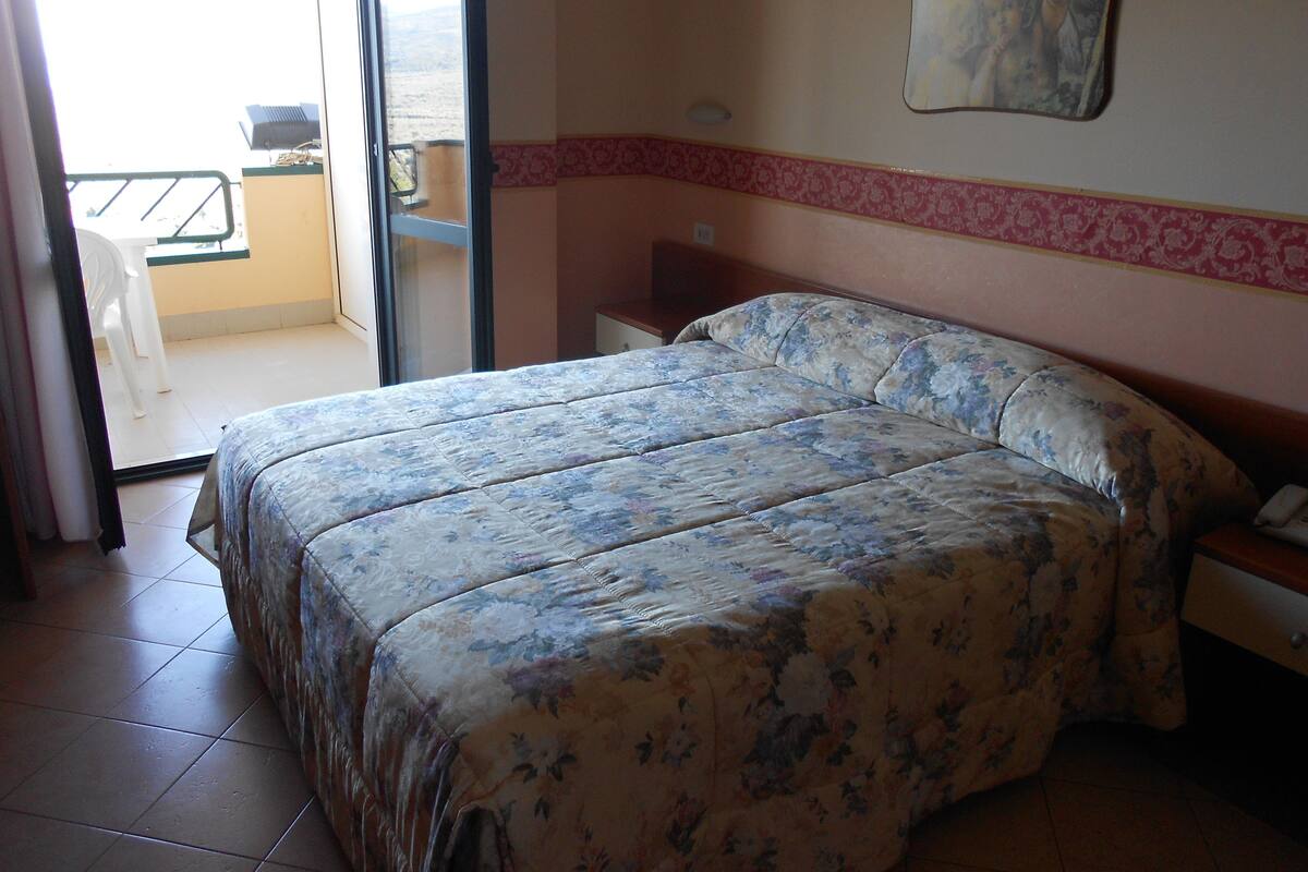 Un dormitorio con edredón estampado de flores, una mesita de noche, una silla y vistas al balcón exterior.