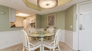 A dining area with a glass table surrounded by wicker chairs, a white door, and a kitchen in the background. - Oceanfront Condominium with Great Amenities (St Simons Island)