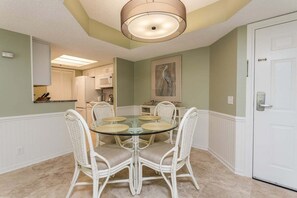 A dining area with a glass table surrounded by wicker chairs, a white door, and a kitchen in the background. - Oceanfront Condominium with Great Amenities (St Simons Island)