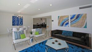 Un salon moderne avec un canapé blanc, une table basse en verre et un tapis bleu. On y trouve également un coin repas avec des chaises blanches et une table blanche. Au fond de la pièce, il y a une cuisine.