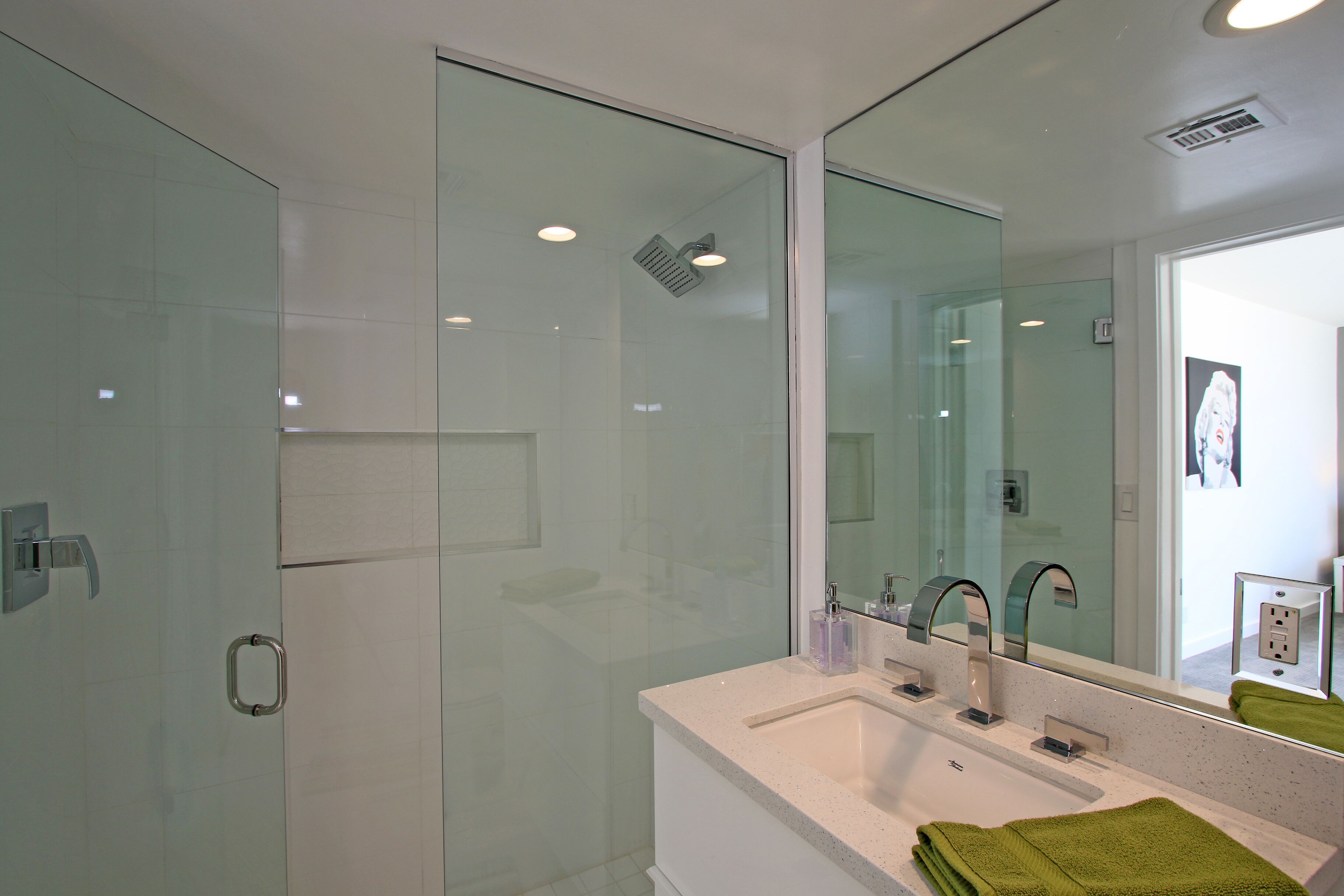 A modern bathroom with a glass shower enclosure, a white sink with a mirror, and a green towel on the counter.