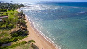 Une vue aérienne d’une plage aux eaux d’un bleu limpide, avec un rivage sablonneux et quelques personnes se promenant le long de la plage.