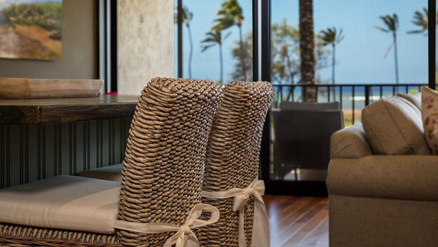 Un salon moderne doté d’une grande fenêtre donnant sur des palmiers et l’océan. On y trouve des fauteuils en osier avec des coussins blancs et un canapé agrémenté d’un coussin à motifs floraux.