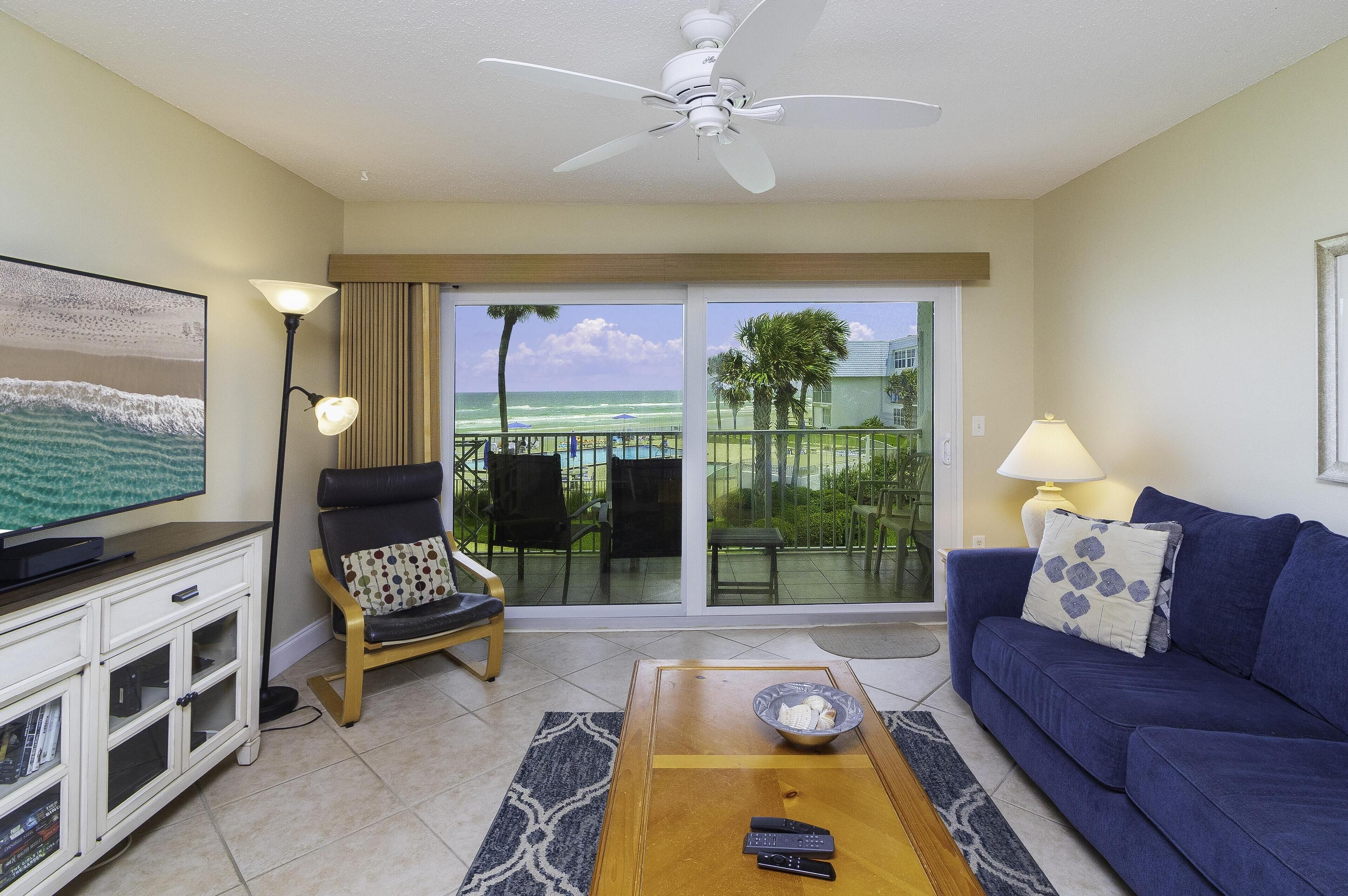 A living room with a TV, a sofa, a coffee table, a chair, and a balcony with a view of the ocean.