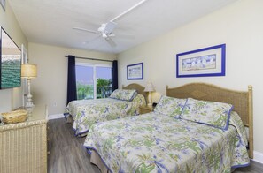 Two single beds with floral-patterned bedding, a ceiling fan, and a large window offering a view of the outdoors.