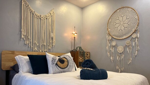 Un lit bien fait, avec une literie blanche et une tête de lit en bois, une décoration murale en macramé, un attrape-rêves et une lampe de chevet.