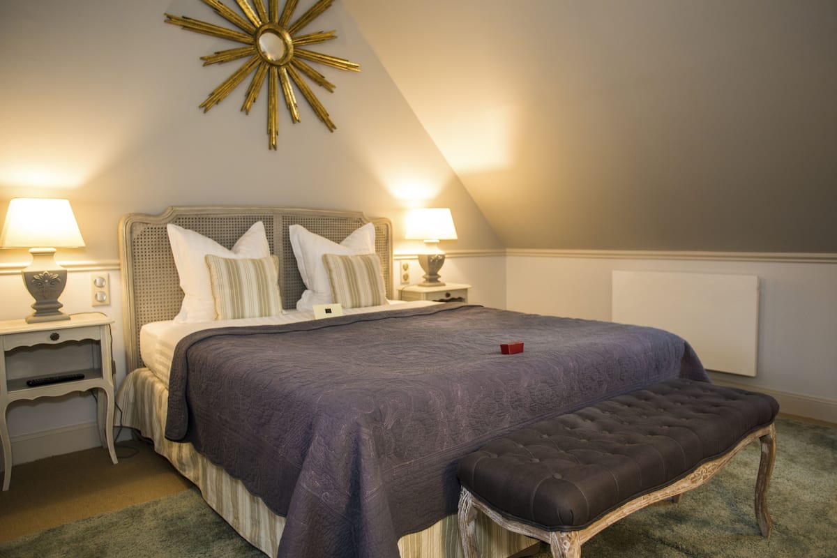 Quarto com uma cama grande, duas mesas de cabeceira, um banco, um objeto decorativo na parede e um radiador.