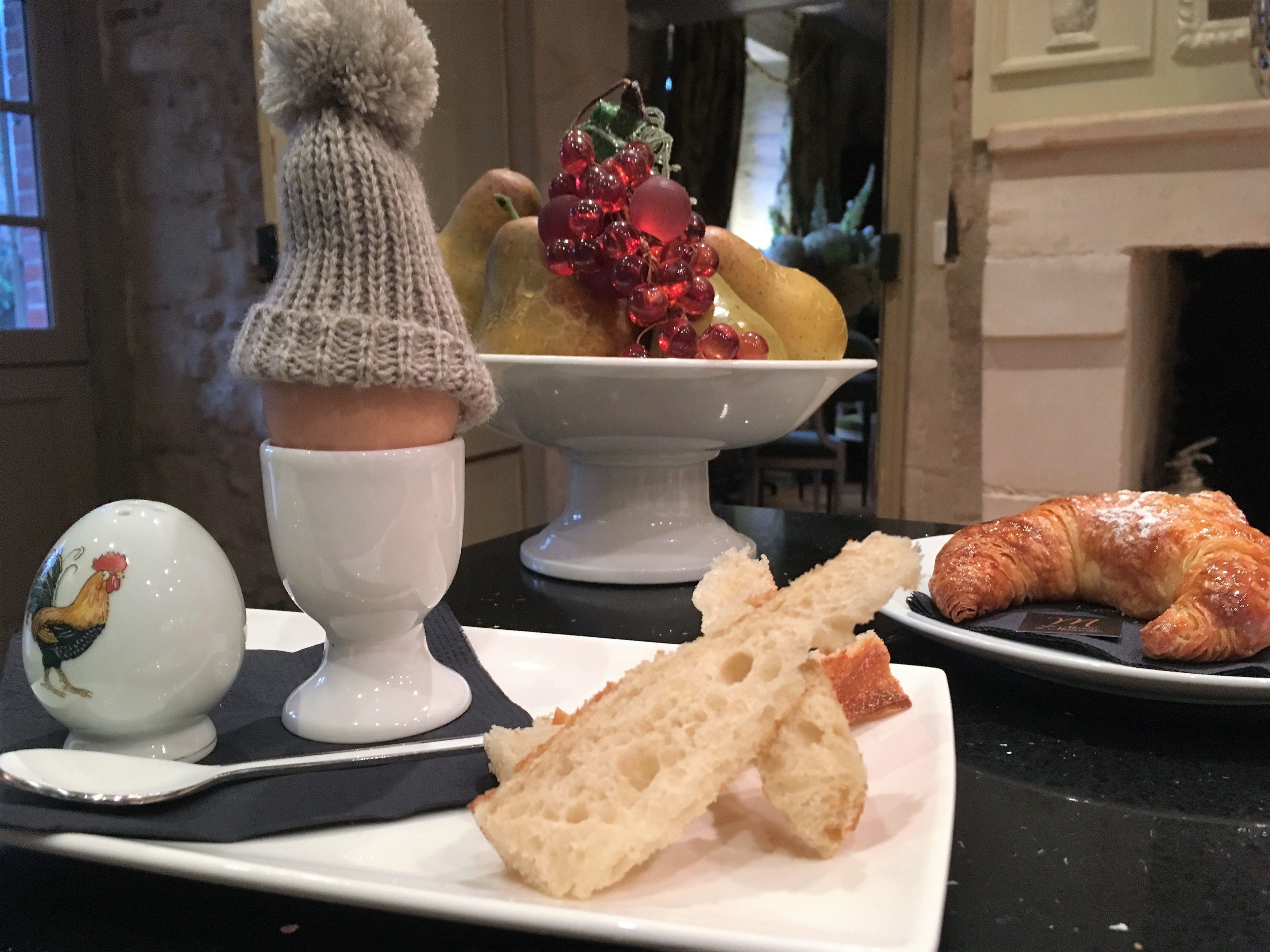 Une table mise pour le petit-déjeuner avec un croissant, du pain, un coq en céramique et un bol de fruits.