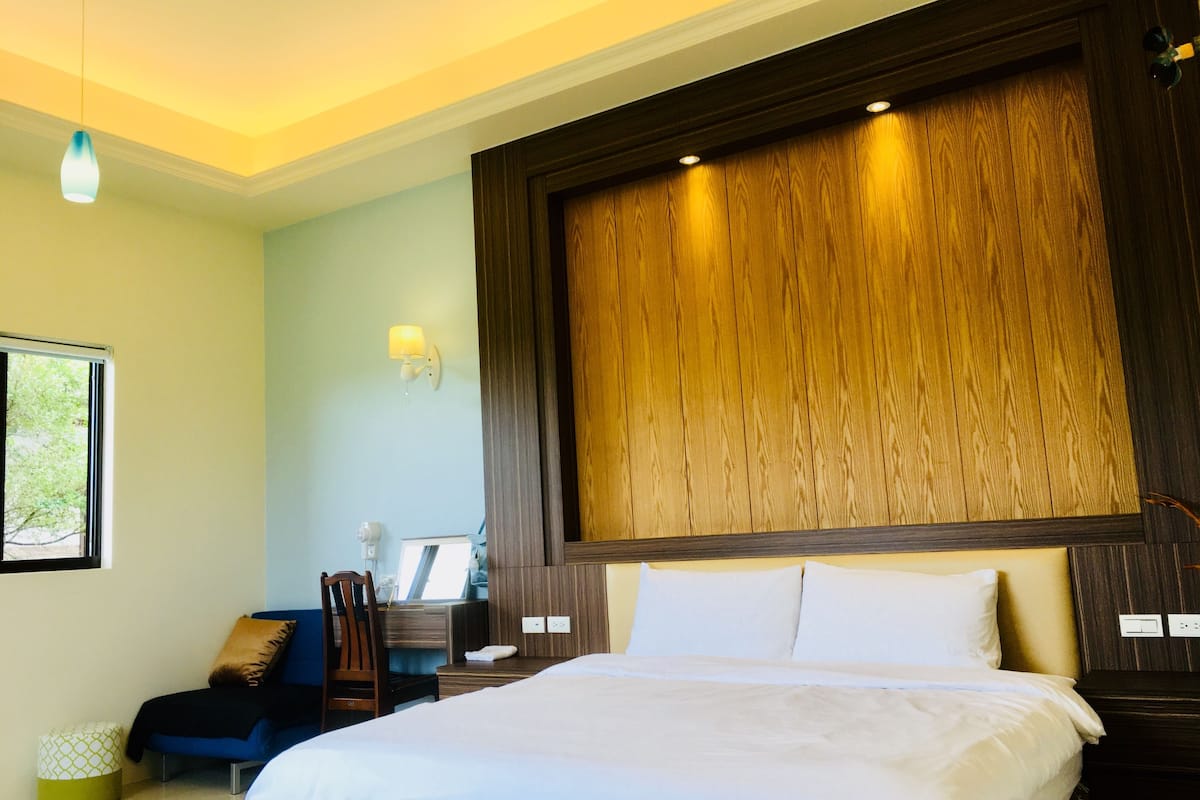 A hotel room with a large bed, a desk, a chair, and a wall-mounted lamp.