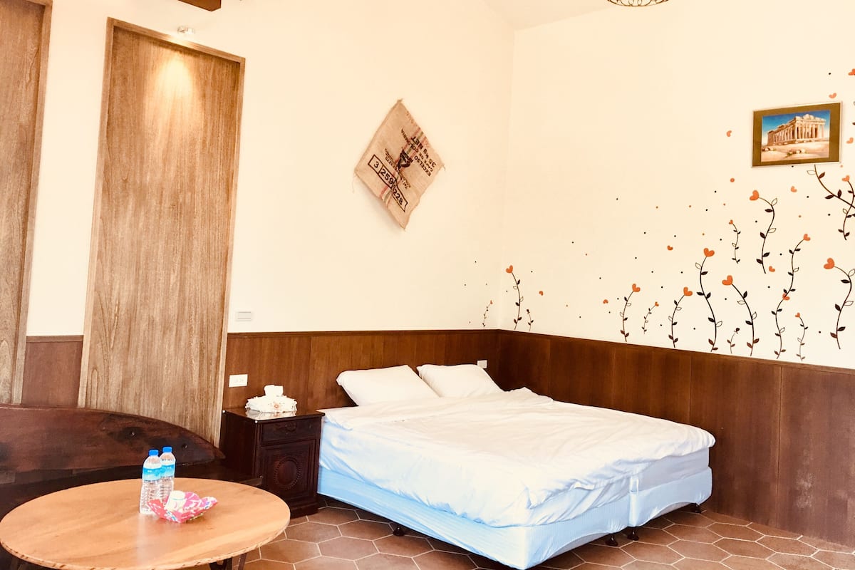 A hotel room with a bed, a wooden headboard, a wall clock, a framed picture, and a small table with bottles.