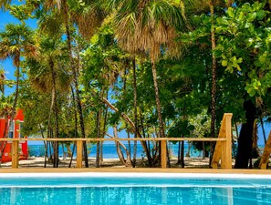 A pool area with a wooden deck, surrounded by palm trees and a beach view.