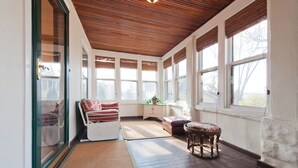 A sunlit room with a wooden ceiling, large windows, a white sofa, a small table, and a wicker stool.