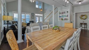 Un vaste espace dédié à la salle à manger, avec une table en bois et des chaises blanches, un escalier menant à un autre niveau, et une bibliothèque contenant divers objets.