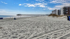 Une plage avec une jetée qui s’avance dans l’océan, un terrain sablonneux et un bâtiment à plusieurs étages situé sur la droite.