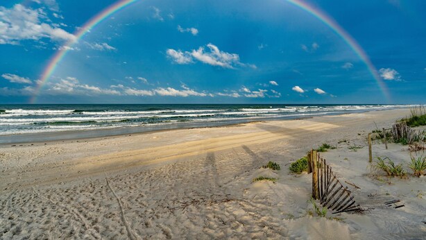 Une plage avec un arc-en-ciel, des dunes de sable et des vagues de l’océan.
