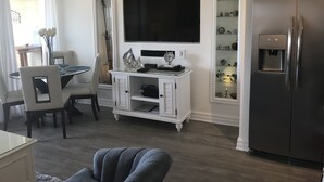 A modern kitchen with a built-in refrigerator, a dining area with a white table and chairs, and a flat-screen TV mounted above the counter.