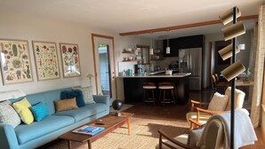 Een woonkamer met een bank, salontafel en fauteuils, een keuken met een centraal eiland en barstoelen, en ingelijste botanische prints aan de muur.