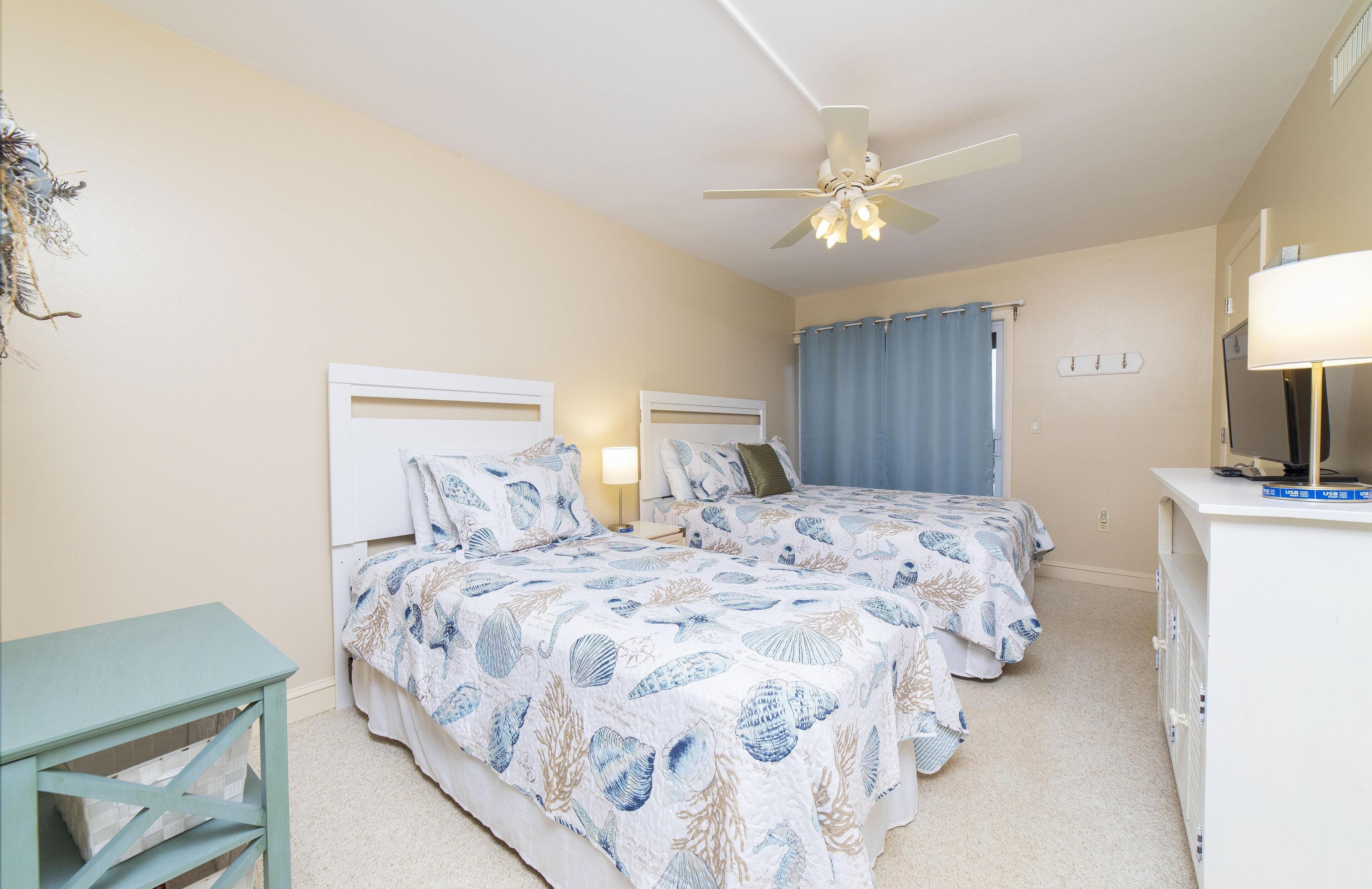 A bedroom with two beds, a ceiling fan, a desk with a lamp, and a television.