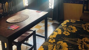 A room with a wooden table, a woven placemat, a bed with a floral bedspread, a window with metal bars, and a curtain.