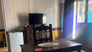 A room with a wooden dining table, a television on a stand, a small refrigerator, and a patterned bedspread.