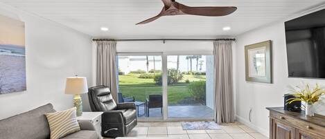 A living room with a ceiling fan, a sofa, a chair, a TV, and a view of a lawn through glass doors.