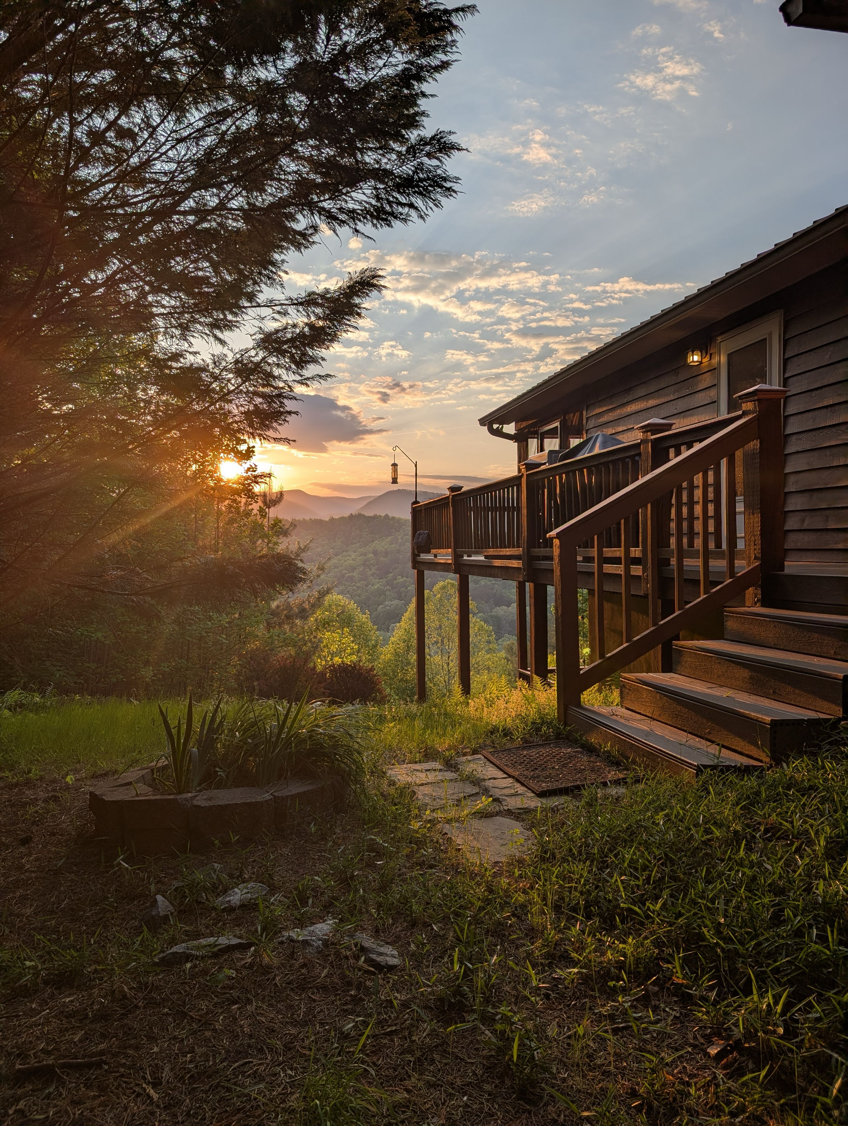 Eine Holzterrasse mit einer Treppe, die zu einem Haus führt, umgeben von viel Grün und einer Berglandschaft im Hintergrund.