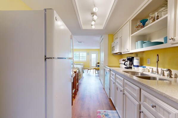 A kitchen with white cabinets, a double sink, and a microwave. There is a dining area in the background with a table and chairs. - Studio Condo in Town, Near Beach, Shared Pool and Hot Tub (Port Aransas)