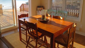Un coin repas avec une table et des chaises en bois, un pichet en verre et une vue sur la plage par une fenêtre munie de stores.