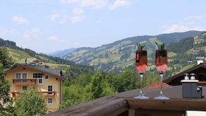 A view of a mountain landscape with houses and a balcony with two glasses of wine. - Apartment Hotel Panorama (Wagrain)