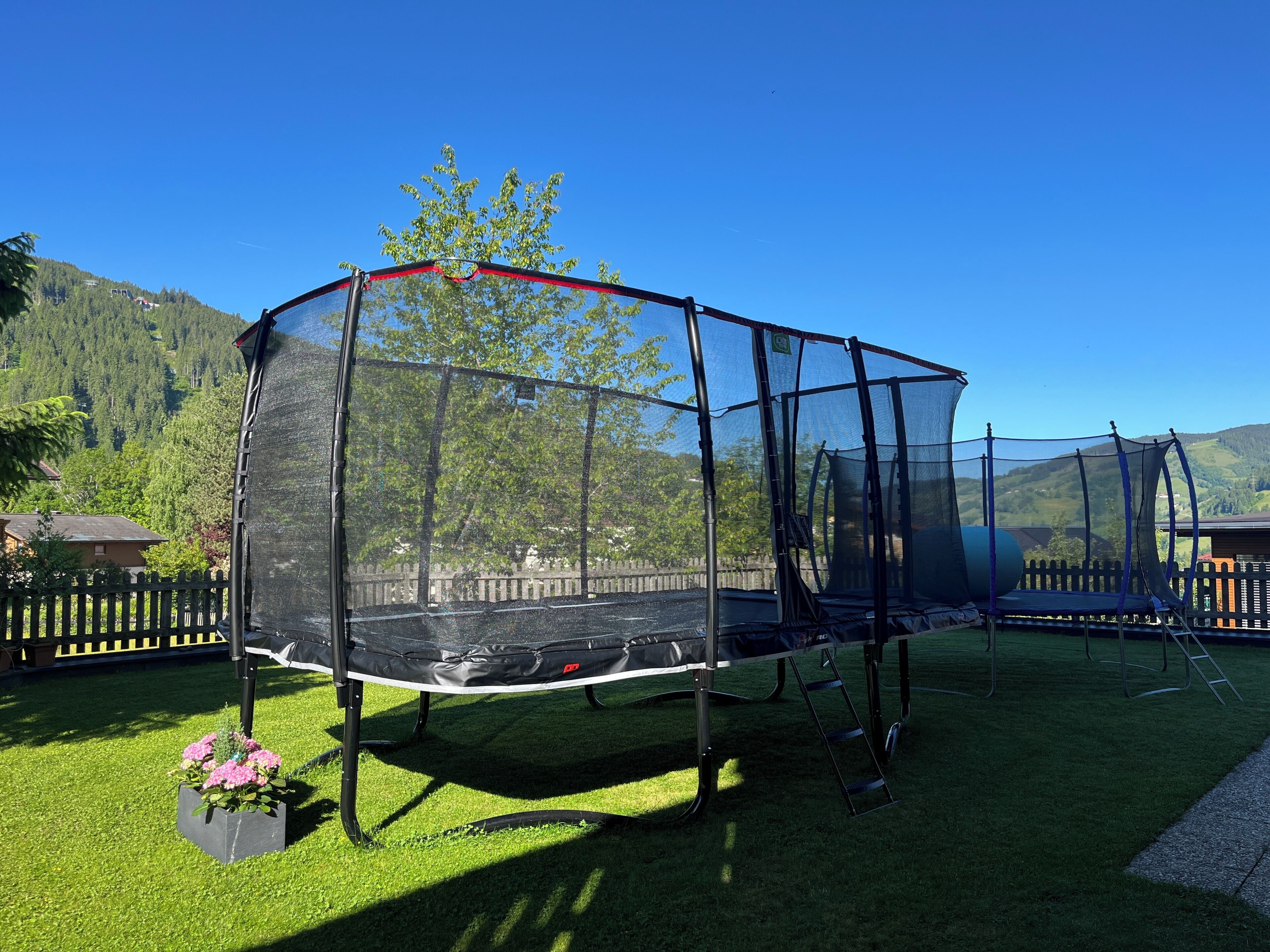 Un trampolín con red de seguridad, rodeado por una valla de madera, sobre un césped con montañas al fondo.