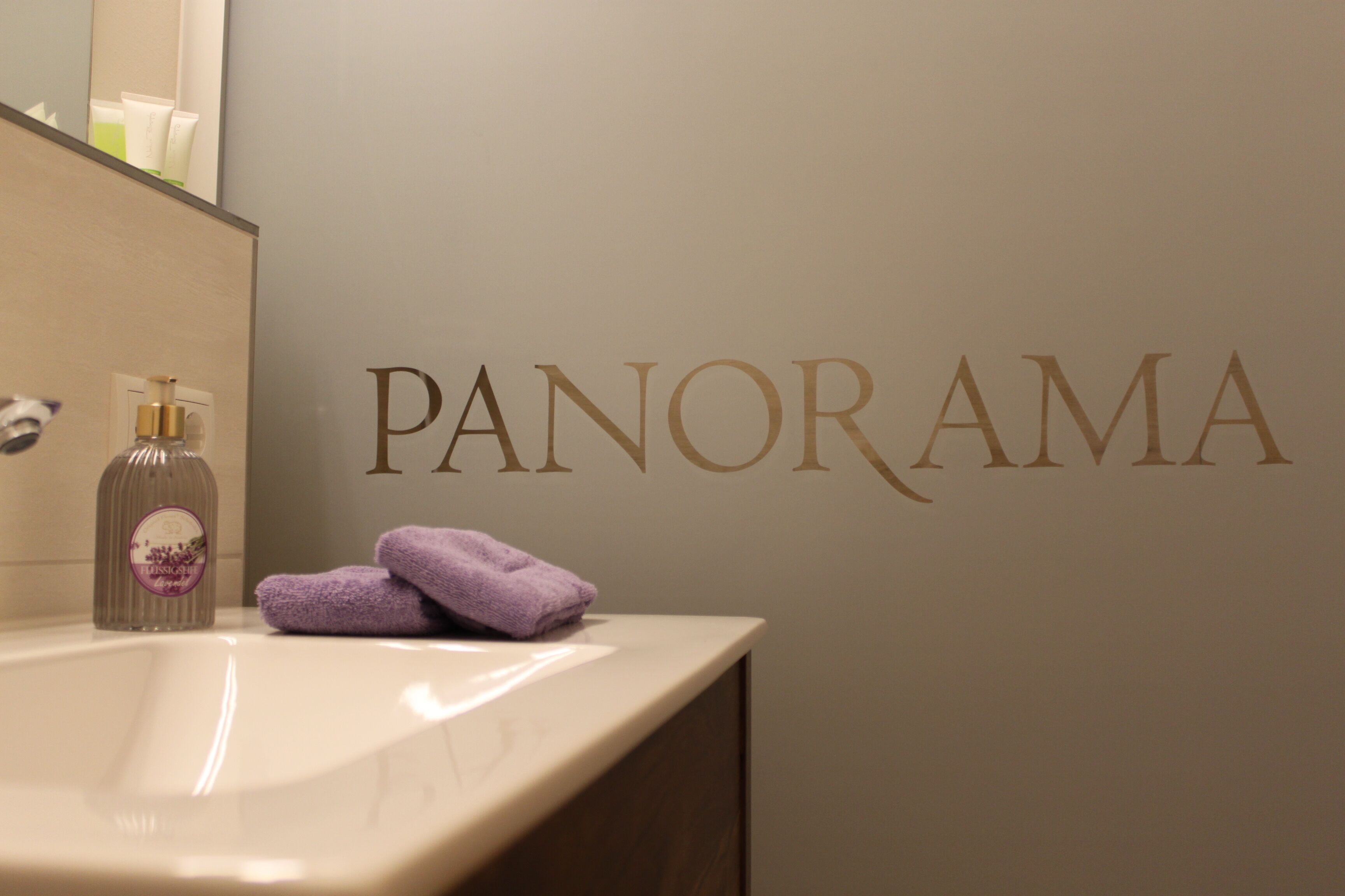A bathroom with a sink, a bottle of lotion, and two towels. The wall has the word "PANORAMA" written on it.