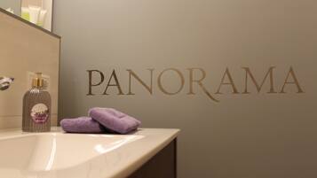 A bathroom with a sink, a bottle of lotion, and two towels. The wall has the word "PANORAMA" written on it.