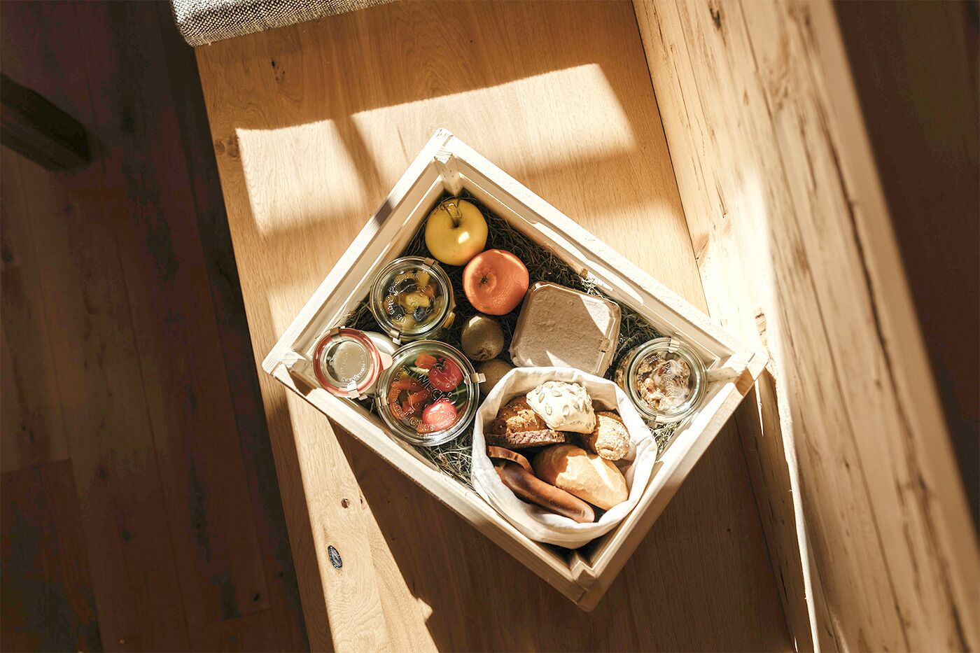 A wooden box containing various food items such as bread, tomatoes, and jars, placed on a wooden surface.