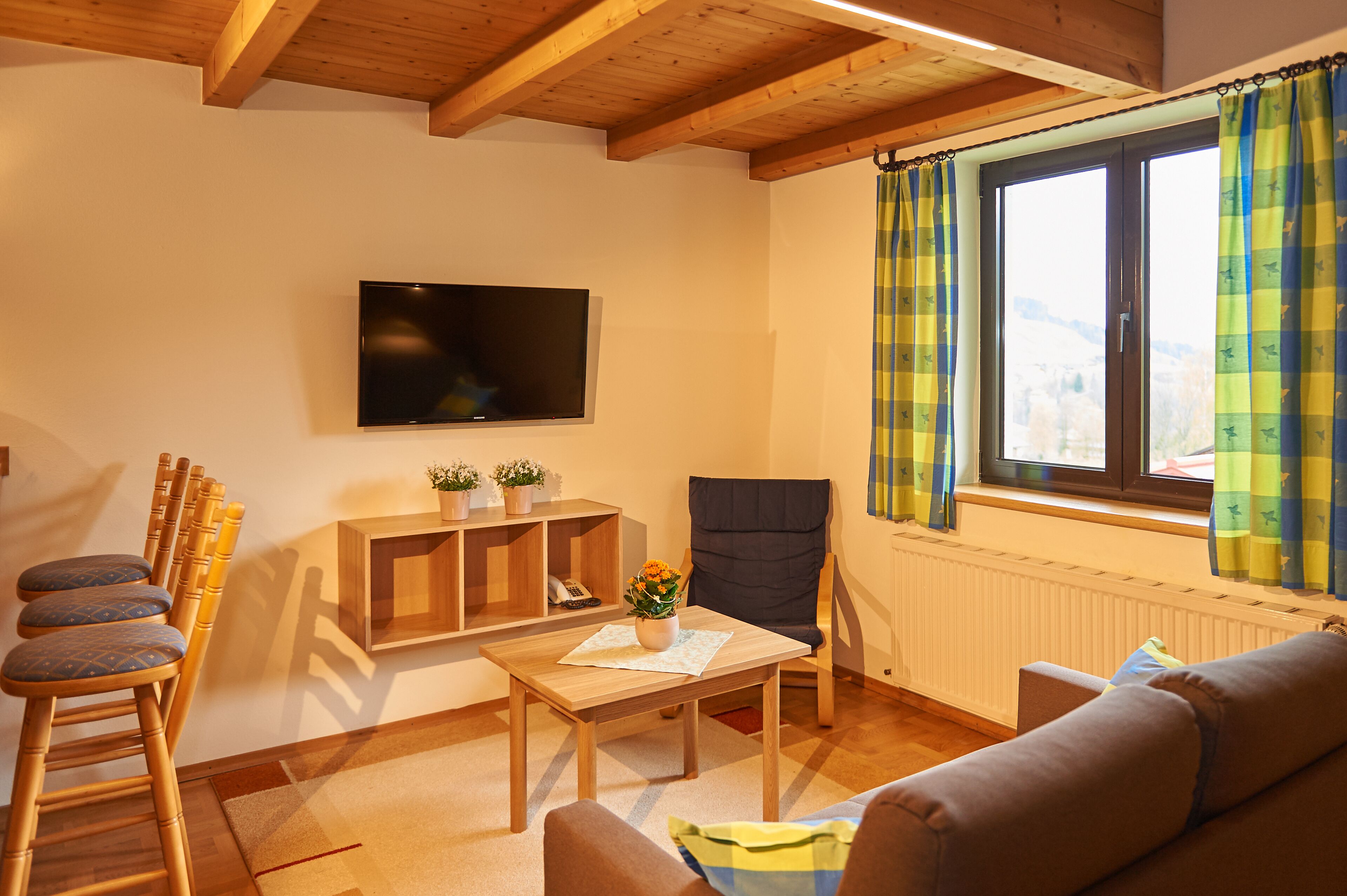 A cozy living room with a wooden ceiling, a flat-screen TV, a sofa, a chair, a table with a vase of flowers, and a window with curtains.