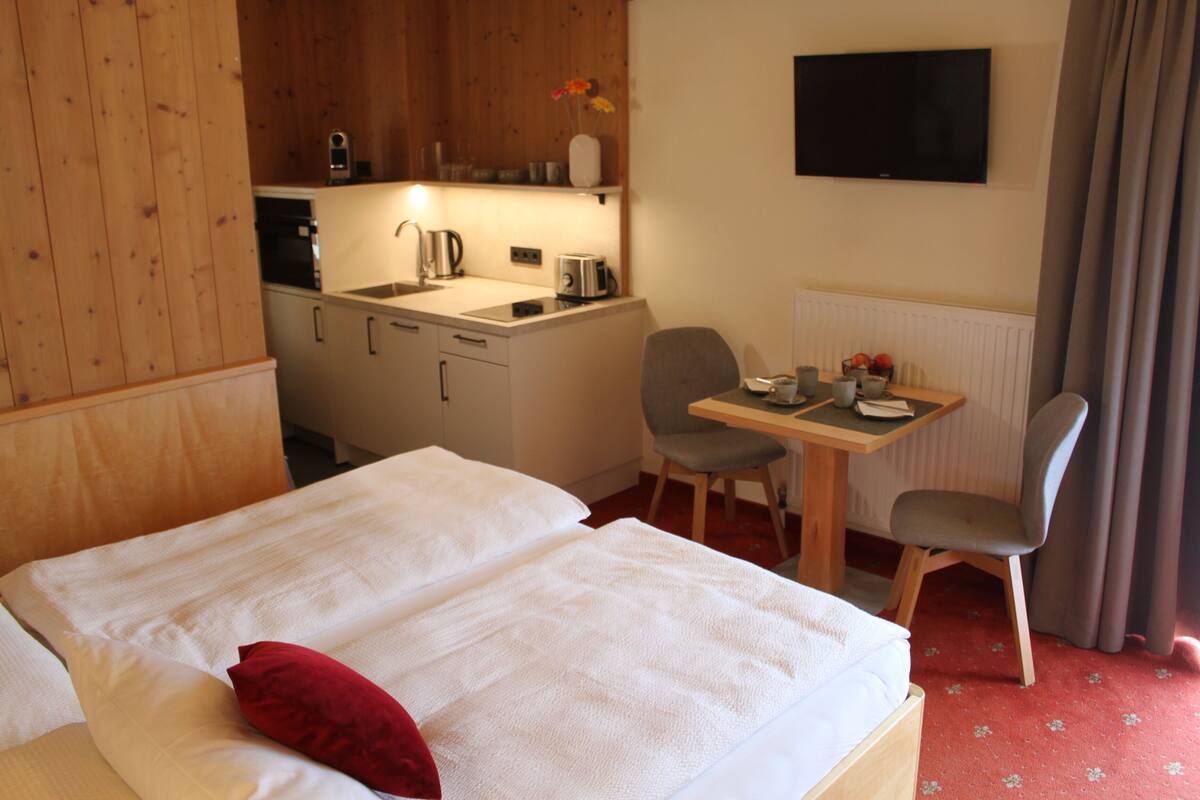 Hotellihuone, jossa on sänky, työpöytä ja kaksi tuolia, pieni keittiö sekä seinälle asennettu televisio.