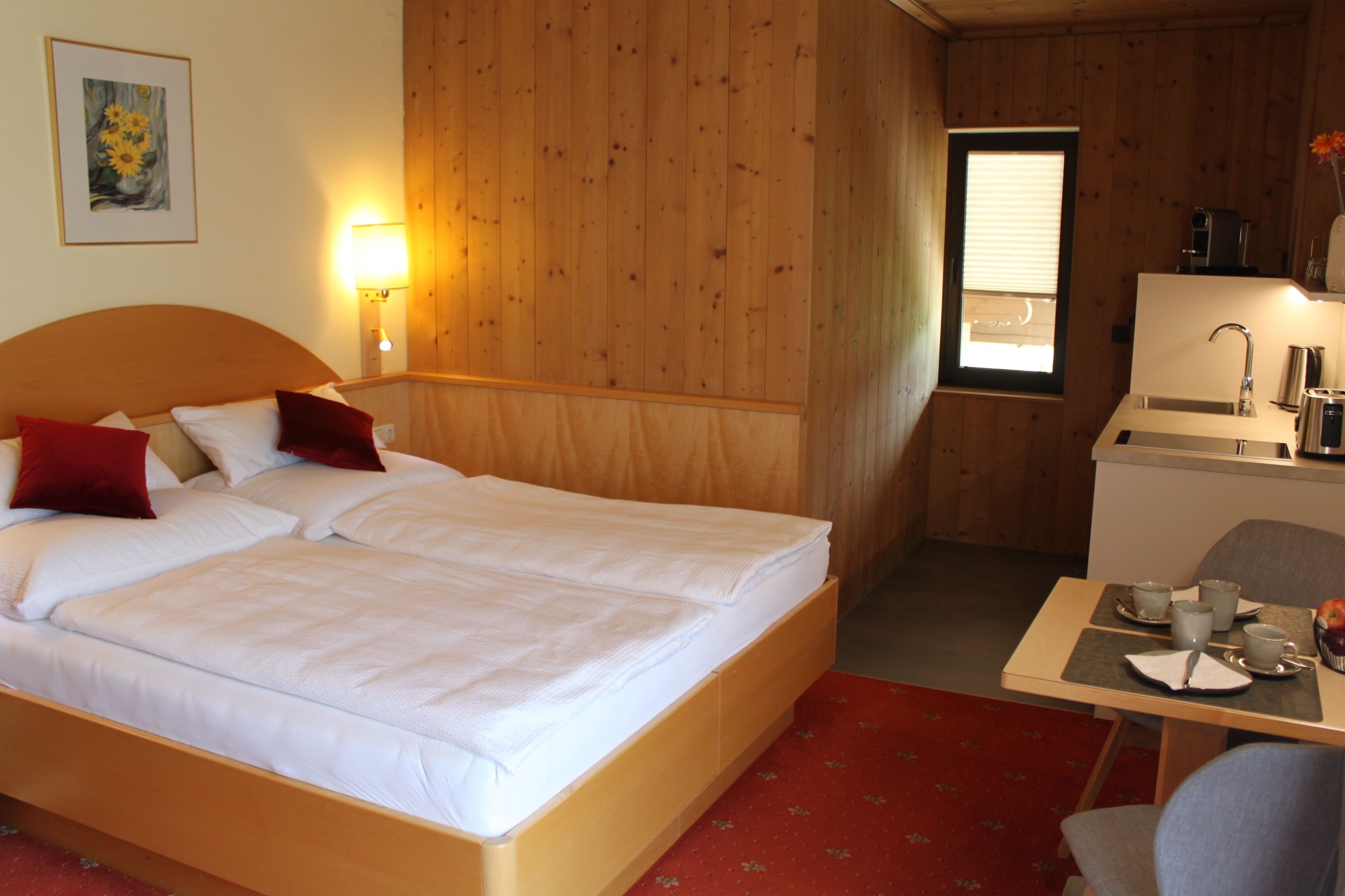 A double bed with white linens, a wooden headboard, a red pillow, a bedside lamp, a small table with cups and a fruit bowl, and a window with blinds.