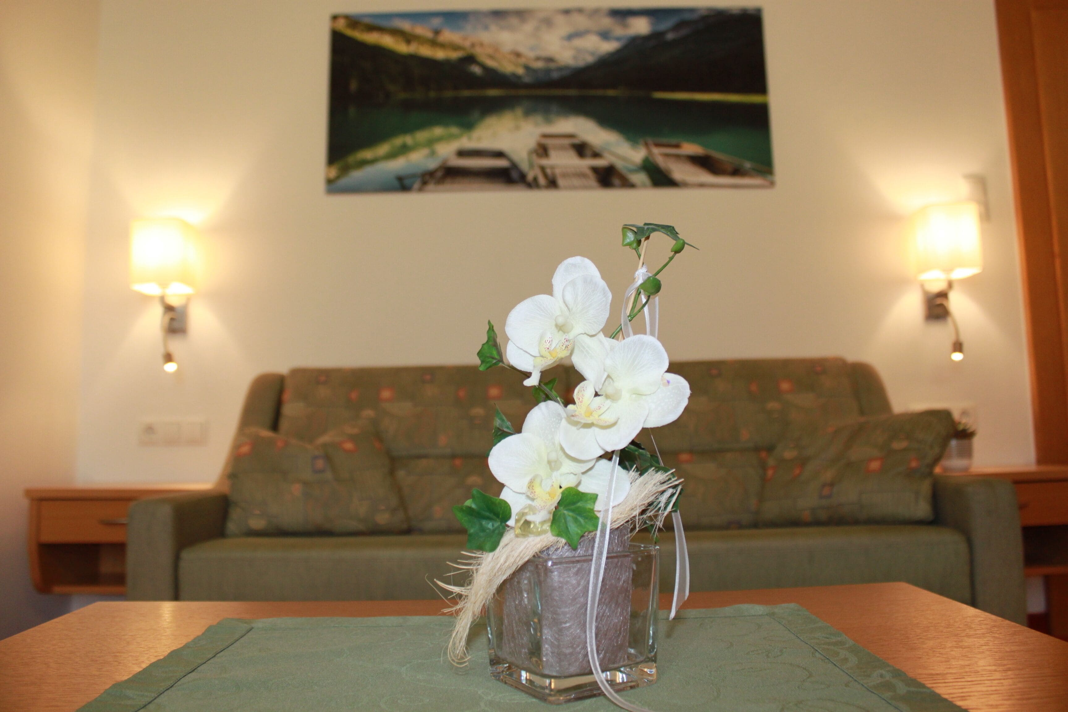 Una habitación con un sofá verde, una mesita con un florero de flores blancas y un cuadro de un lago en la pared.