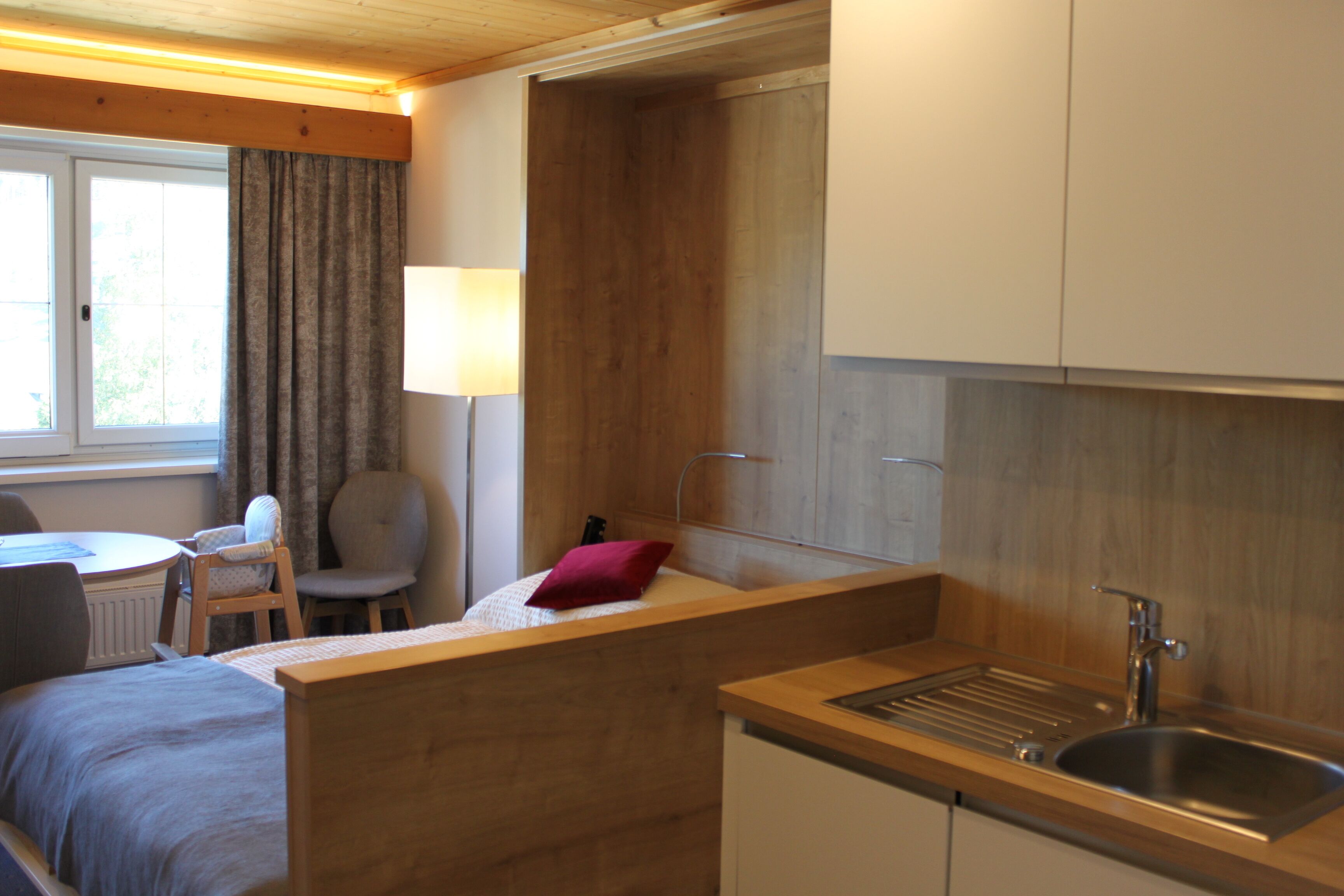 A compact room with a bed, a small desk, a chair, a lamp, and a kitchenette with a sink.