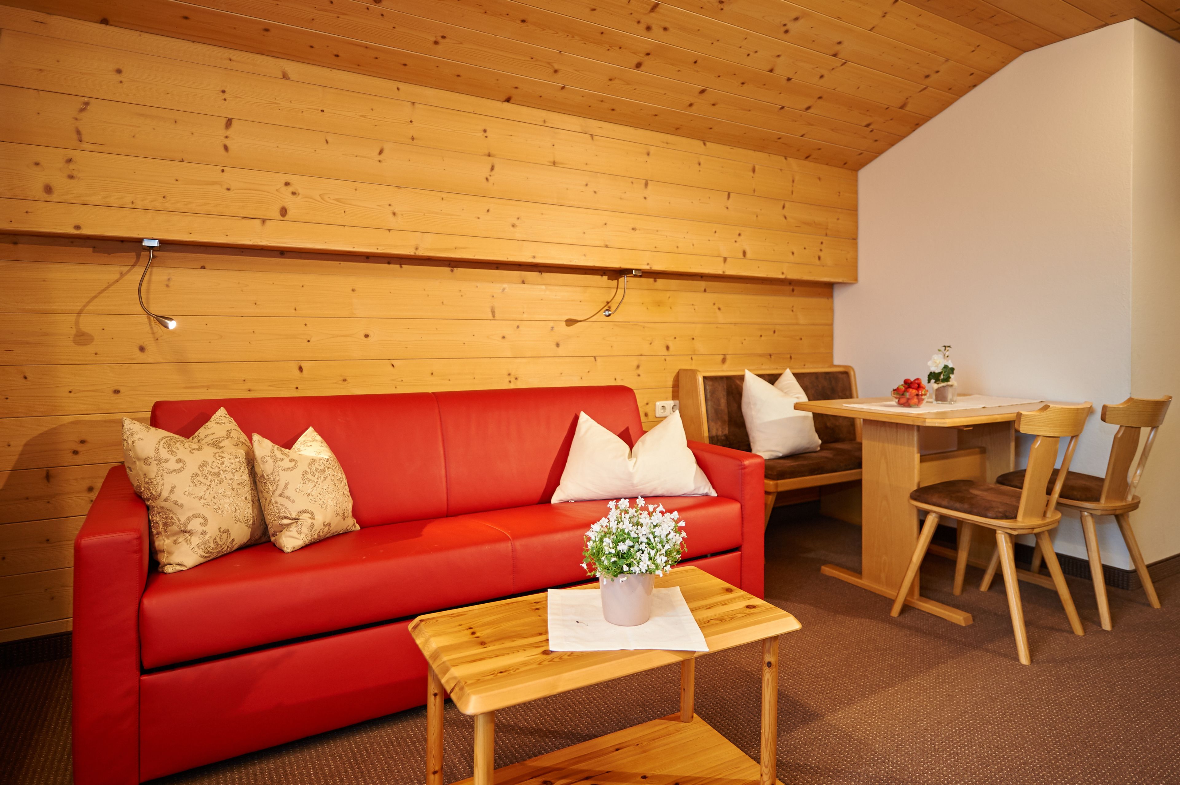 Een rode bank met patroon kussens, een houten tafel met een plant in pot, en houten stoelen in een kamer met houten wanden en plafond.
