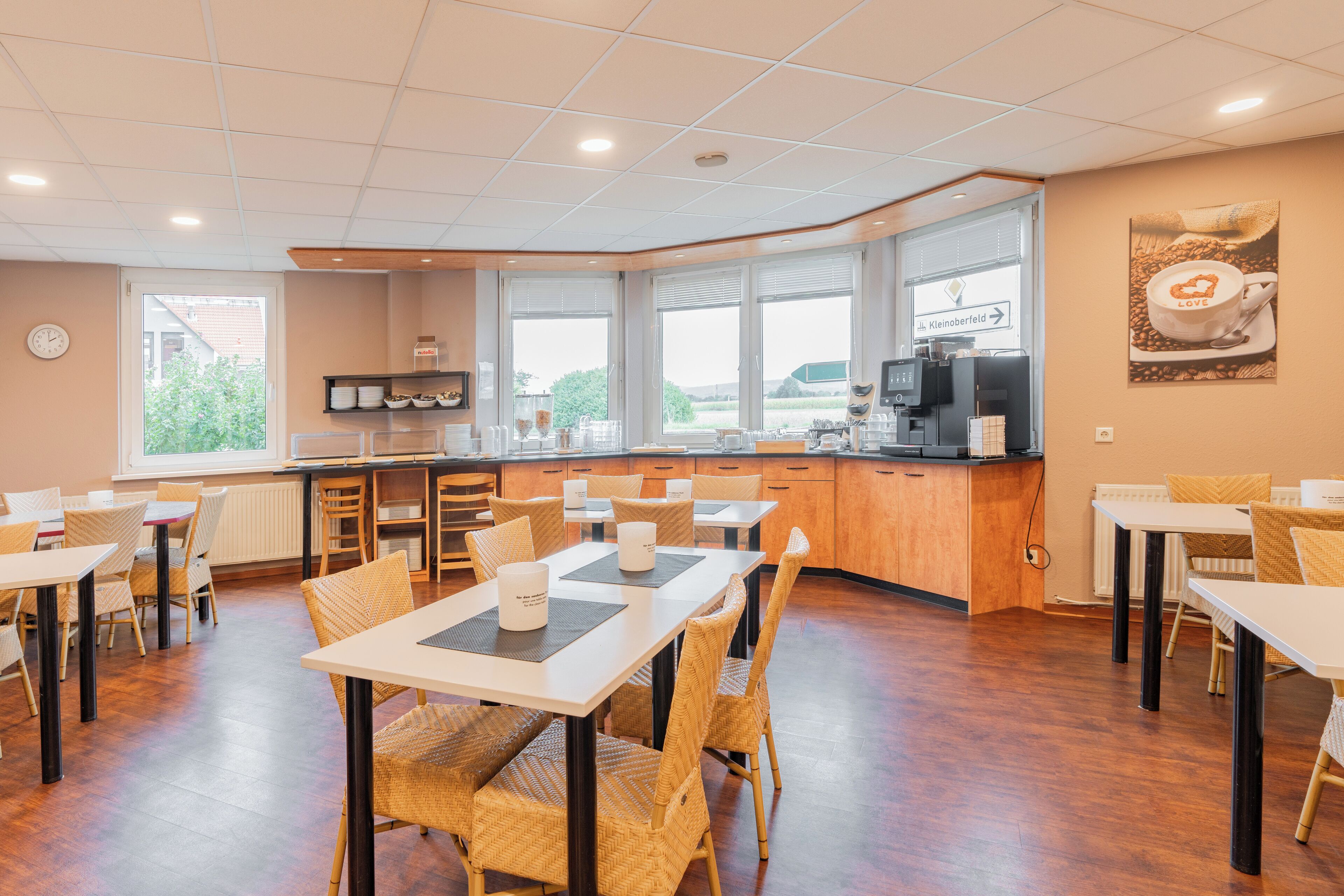 Un espace repas moderne avec des tables et des chaises en bois, un comptoir de cuisine et de grandes fenêtres donnant sur l’extérieur.