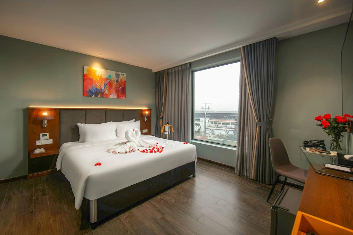 Ett hotellrum med en stor säng, ett skrivbord, en stol, utsikt över staden och en tavla på väggen.